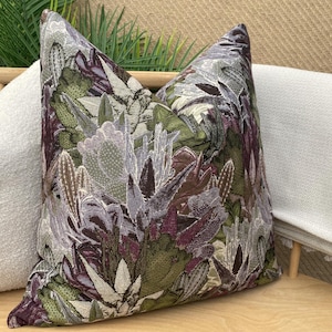 May include: A decorative throw pillow with a floral pattern featuring shades of green, purple, and white. The pillow is made of a textured fabric and has a subtle sheen.