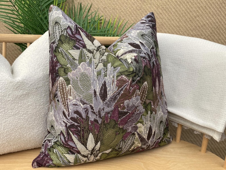 May include: A decorative throw pillow with a floral pattern featuring shades of green, purple, and white. The pillow is made of a textured fabric and has a subtle sheen.