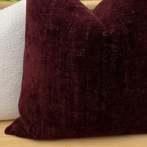 Pu&ograve; includere: Due cuscini decorativi. Uno &egrave; di colore bordeaux scuro con un aspetto strutturato e vellutato. L'altro cuscino &egrave; bianco con una superficie strutturata e nodosa. I cuscini sono probabilmente per l'arredamento della casa.