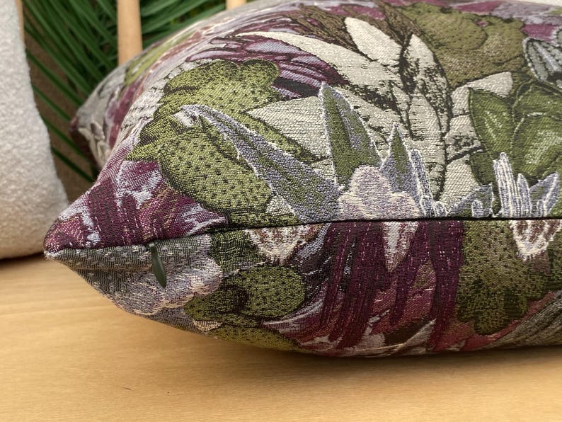 May include: A close-up of a decorative pillow with a floral pattern. The pillow is made of a textured fabric with a variety of colors, including green, purple, and white.