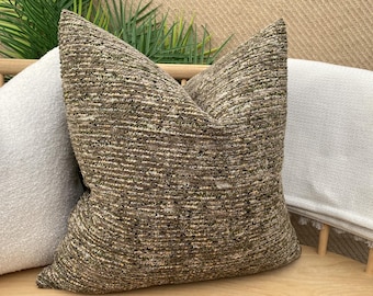 Può includere: Un cuscino decorativo di colore marrone e beige con una texture e un leggero bagliore. Il cuscino è su un divano grigio chiaro.