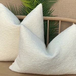 May include: Two white fluffy throw pillows on a wooden bench with green plants in the background.