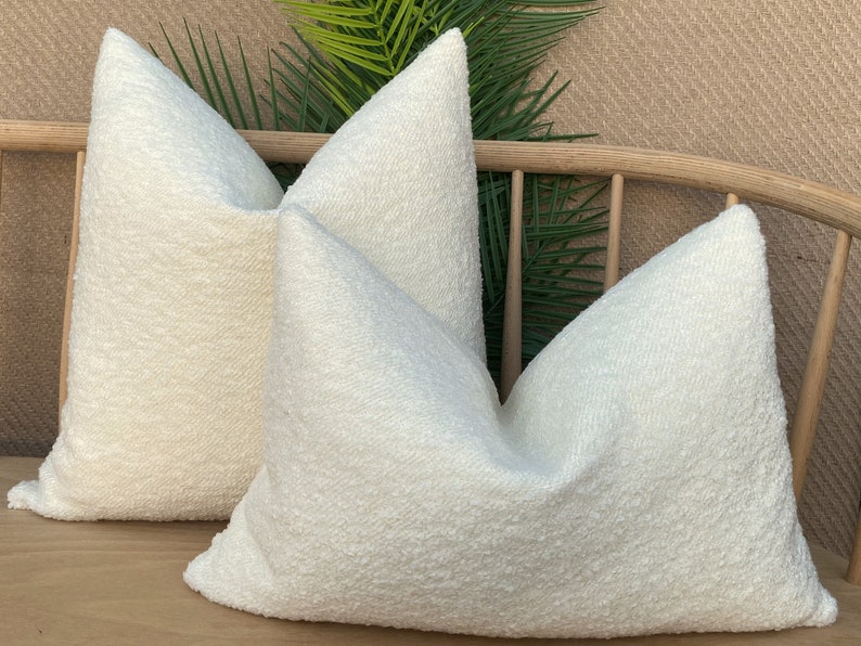 May include: Two white fluffy throw pillows on a wooden bench with green plants in the background.