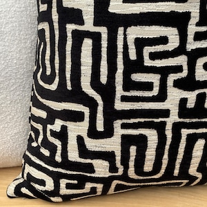 Puede incluir: Primer plano de un cojín decorativo con un patrón geométrico negro y blanquecino. El diseño presenta un motivo laberíntico. El cojín está hecho de una tela texturizada y forma parte de una colección de decoración del hogar.