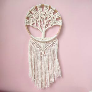 Peut inclure: Une suspension murale en macramé blanche en forme d'arbre avec un cercle en bois. L'arbre a un design détaillé avec des nœuds complexes et une frange de fil blanc.