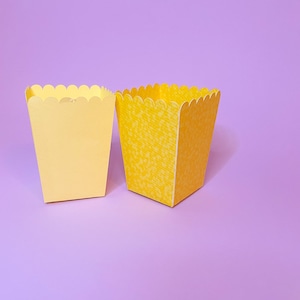 Peut inclure: Deux boîtes en papier jaunes avec des bords festonnés. La boîte de gauche est jaune unie, tandis que la boîte de droite a un motif subtil. Les deux boîtes sont posées sur une surface violette.
