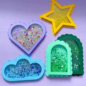 Puede incluir: Cuatro marcos de papel con bordes festoneados, cada uno lleno de purpurina. Los marcos tienen forma de corazón, estrella, nube y arco. La purpurina es de diferentes colores, incluyendo azul, verde y dorado.