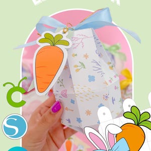 Puede incluir: Una caja con forma de huevo de Pascua blanca y azul con un diseño de zanahoria. La caja está decorada con flores, gotas de lluvia y una cinta azul. La caja está siendo sostenida por una mano con esmalte de uñas rosa. La caja está etiquetada como "Easter Box".