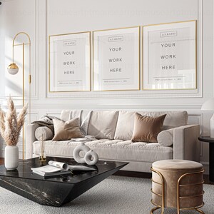 May include: Three gold framed posters with the text "YOUR WORK HERE" on a white wall above a white sofa with brown pillows. A black marble coffee table with a magazine and two decorative rings sits in front of the sofa. A gold floor lamp with a round light fixture is in the corner of the room. A beige ottoman with a gold metal frame sits in front of the sofa.