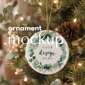 May include: A round white ceramic ornament featuring a green watercolor eucalyptus leaf wreath design. The center contains the black text YOUR design HERE. The ornament hangs from a gold string and is displayed against a pine tree background.