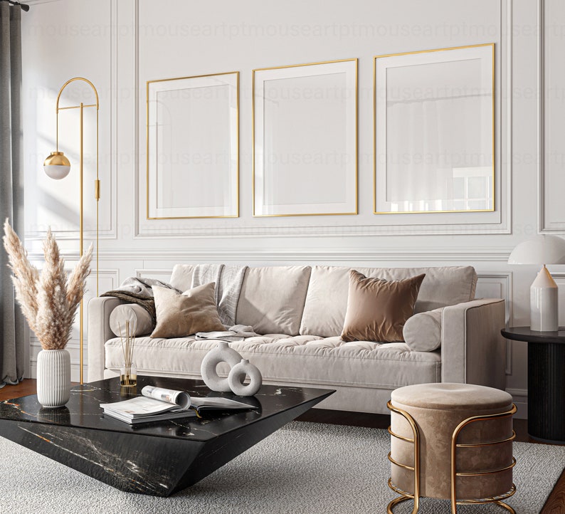 May include: A living room interior design with a white sofa, three gold framed pictures, a black coffee table, a gold floor lamp, a beige ottoman, and a dried pampas grass in a white vase.