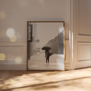May include: A black and white photograph of a person walking down a city street with an umbrella. The person is wearing a coat and carrying a bag. The photo is framed in a wooden frame.