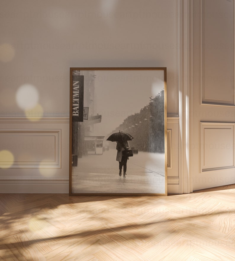 May include: A black and white photograph of a person walking down a city street with an umbrella. The person is wearing a coat and carrying a bag. The photo is framed in a wooden frame.