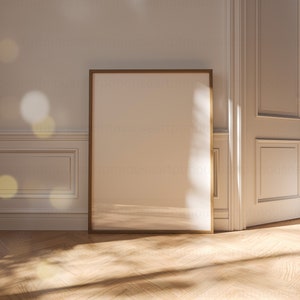 May include: A large, empty, light brown picture frame with a thin, gold border sits on a light wood floor. The frame is leaning against a white wall with a white door to the right.
