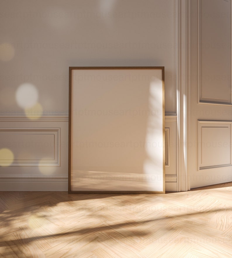 May include: A large, empty, light brown picture frame with a thin, gold border sits on a light wood floor. The frame is leaning against a white wall with a white door to the right.