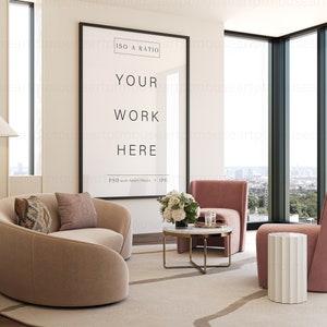 May include: A modern living room interior with a large framed poster that says "ISO A RATIO YOUR WORK HERE PSD with Smart Object + JPG". The poster is hanging on a white wall. There is a beige curved sofa with two pillows, a pink armchair, a white side table, and a coffee table with a vase of white flowers. The room has large windows with a view of a city skyline.