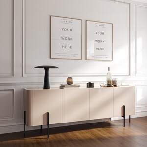 May include: A light beige sideboard with four doors and black metal legs. The sideboard is decorated with a black lamp, two framed posters with the text "YOUR WORK HERE", a glass vase with a brown glass object inside, and a gold tray with a bottle and glasses.