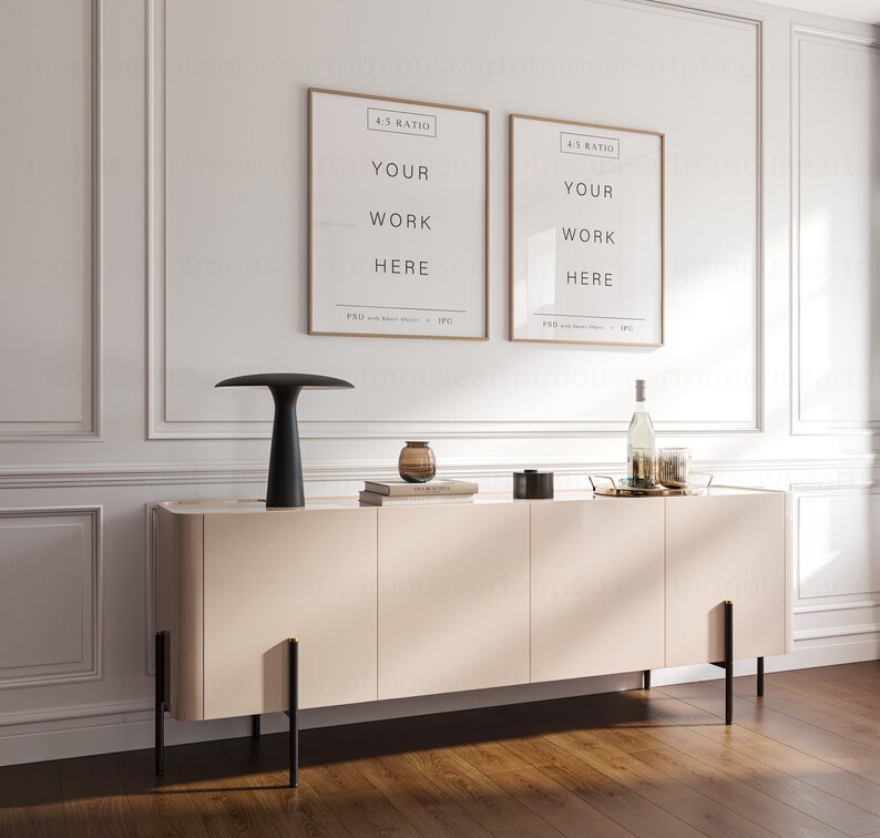May include: A light beige sideboard with four doors and black metal legs. The sideboard is decorated with a black lamp, two framed posters with the text "YOUR WORK HERE", a glass vase with a brown glass object inside, and a gold tray with a bottle and glasses.