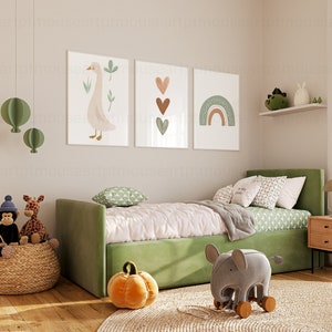 May include: A green upholstered daybed with a white duvet and green patterned pillows. There are three framed prints on the wall above the bed, featuring a goose, hearts, and a rainbow. A stuffed elephant toy sits on the floor in front of the bed.