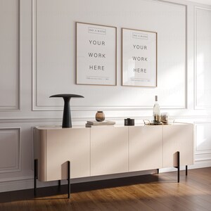 May include: A light beige cabinet with black metal legs and three doors. Two framed posters with the text "ISO A RATIO YOUR WORK HERE" are hanging on the wall above the cabinet. A black lamp with a white lampshade is on the cabinet, along with a few books and a decorative glass vase.