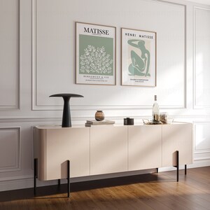 May include: A light beige sideboard with black metal legs and three doors. Two framed posters with the text "MATISSE" and "HENRI MATISSE" are hanging on the wall above the sideboard. A black lamp with a white shade is on the sideboard, along with a clear glass vase with a brown liquid inside.