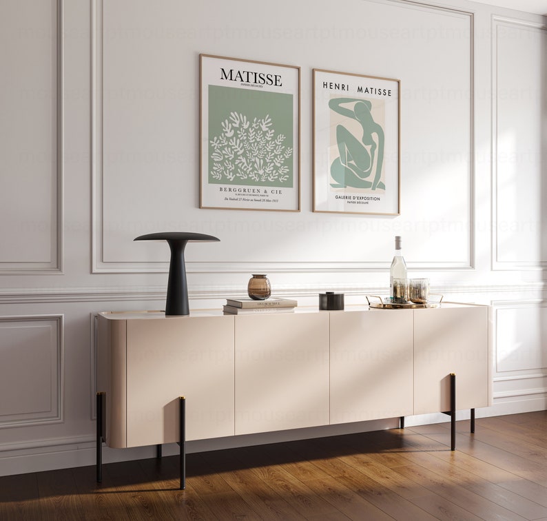 May include: A light beige sideboard with black metal legs and three doors. Two framed posters with the text "MATISSE" and "HENRI MATISSE" are hanging on the wall above the sideboard. A black lamp with a white shade is on the sideboard, along with a clear glass vase with a brown liquid inside.