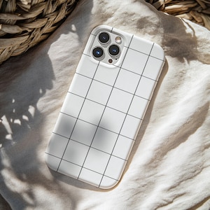 May include: A white phone case with a black grid pattern. The case has a camera cutout for a multi-lens camera system. The case is resting on a beige fabric surface, with a woven basket in the background.