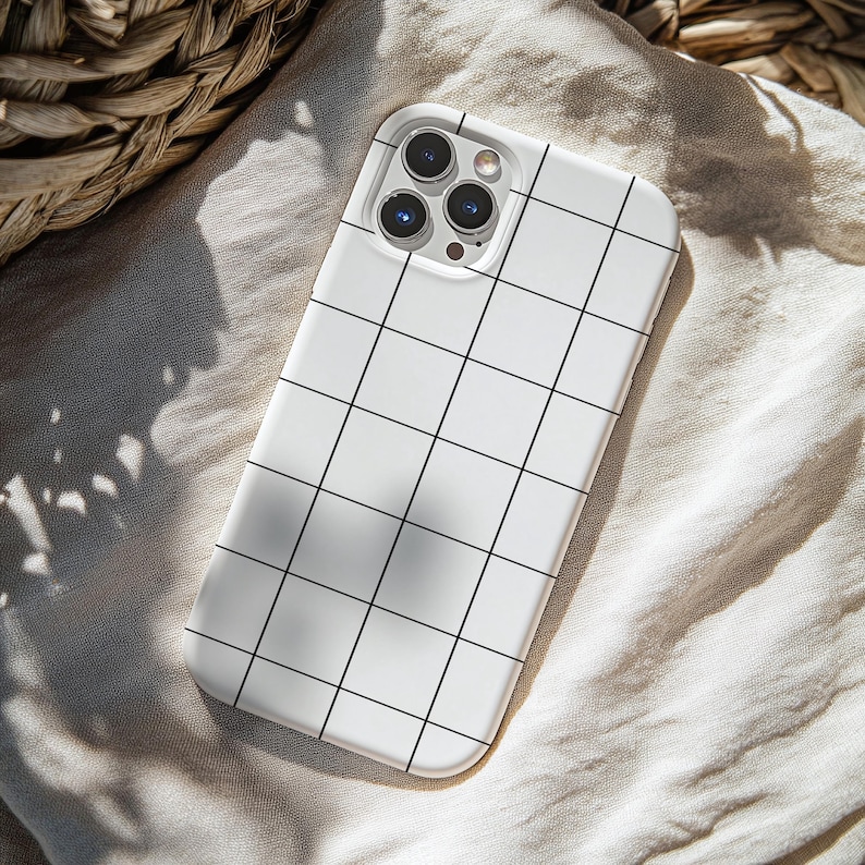 May include: A white phone case with a black grid pattern. The case has a camera cutout for a multi-lens camera system. The case is resting on a beige fabric surface, with a woven basket in the background.