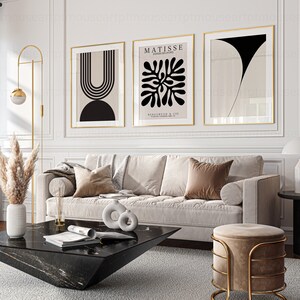 May include: Three framed abstract art prints in black and white with gold frames hanging on a white wall above a beige sofa with brown pillows. A black marble coffee table with a magazine on top sits in front of the sofa. A gold floor lamp with a round shade is in the corner of the room. A gold stool with a beige cushion is in the foreground.
