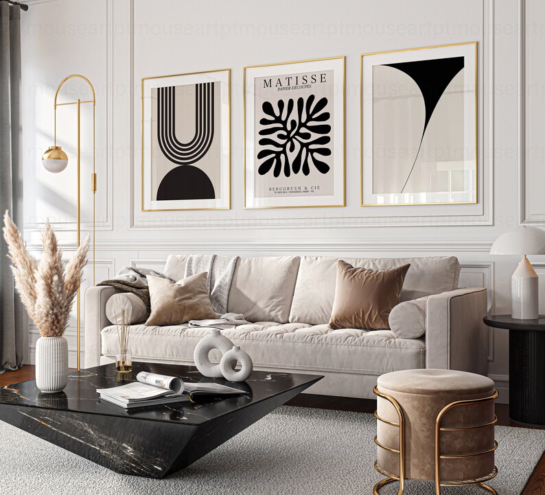 May include: Three framed abstract art prints in black and white with gold frames hanging on a white wall above a beige sofa with brown pillows. A black marble coffee table with a magazine on top sits in front of the sofa. A gold floor lamp with a round shade is in the corner of the room. A gold stool with a beige cushion is in the foreground.