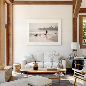 May include: A white sofa with a throw blanket sits in a living room with a wooden coffee table, a vase with dried branches, and a framed photograph of a person walking dogs on a beach.