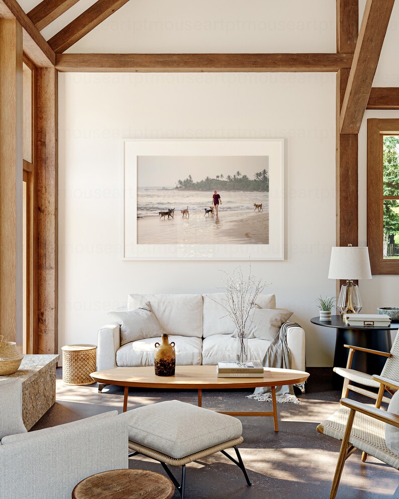 May include: A white sofa with a throw blanket sits in a living room with a wooden coffee table, a vase with dried branches, and a framed photograph of a person walking dogs on a beach.