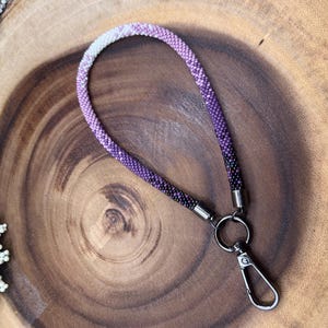 Pode incluir: Um cordão de contas roxo e branco com um fecho giratório prateado. O cordão está sobre uma superfície de madeira.