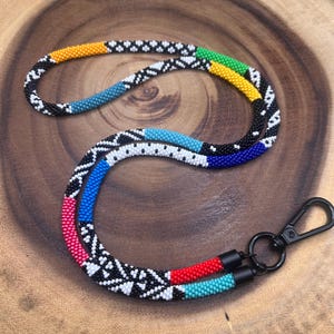 Könnte beinhalten: Ein gefädelter Schlüsselband mit einem schwarzen Verschluss. Das Schlüsselband weist ein sich wiederholendes schwarz-weißes Muster mit Abschnitten aus gelben, grünen, blauen, rosa, roten und türkisfarbenen Perlen auf. Das Schlüsselband wird auf einer Holzoberfläche präsentiert.