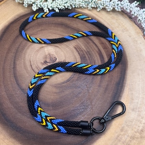 Könnte beinhalten: Schwarzes Perlen-Lanyard mit blauen, gelben und türkisfarbenen Akzenten. Das Lanyard hat ein sich wiederholendes Muster und einen schwarzen Metallverschluss. Das Perlenmuster ist über die gesamte Länge des Lanyards sichtbar.