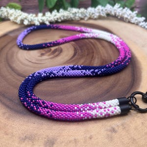 Peut inclure: Un cordon en perles avec un dégradé de perles violettes, roses et blanches. Le cordon est doté de fermoirs métalliques noirs.