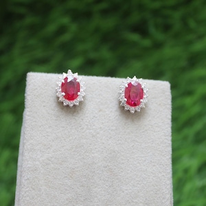 May include: Stud earrings with oval, vibrant red gemstones, each encircled by a halo of smaller, clear stones. The earrings are set in a silver-coloured metal. They are displayed on a neutral surface, with a blurred green backdrop.
