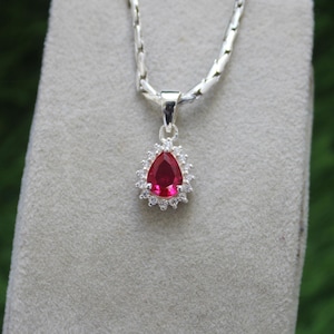 May include: A silver necklace with a teardrop-shaped red gemstone pendant surrounded by a halo of smaller white gemstones.