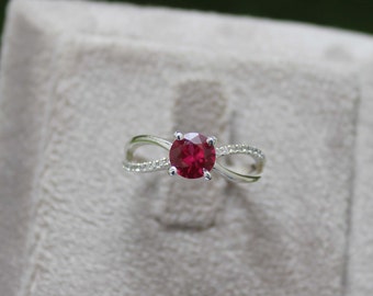 Puede incluir: Un anillo de tono plateado con una piedra central redonda de color rojo vibrante sujeta por cuatro garras. La banda tiene un diseño cruzado con pequeñas piedras claras y brillantes incrustadas a lo largo del brazo dividido, creando un aspecto pulido.