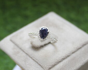 May include: A silver ring featuring a pear-shaped, dark blue gemstone, encircled by a halo of small, clear stones. The ring band is also set with small, clear stones. The ring is presented on a light grey surface.