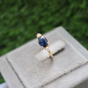 Pode incluir: Um anel de ouro com um safira azul oval e dois diamantes brancos menores. O anel está em uma caixa de veludo cinza.