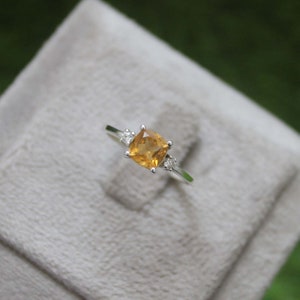 Op de afbeelding: Een zilveren ring met een vierkante, goudgele edelsteen als middelpunt, geflankeerd door twee kleinere, heldere edelstenen. De ring wordt gepresenteerd tegen een zachte, neutrale achtergrond, die het ontwerp en de kleur van het sieraad benadrukt.