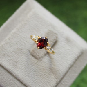 Op de afbeelding: Een gouden ring met een rode ovale edelsteen omringd door kleinere witte edelstenen.