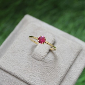 May include: A gold ring featuring a single round, faceted, vibrant red gemstone set in a four-prong setting. The band is thin and polished, showcasing a classic design that highlights the brilliant colour and clarity of the central stone.