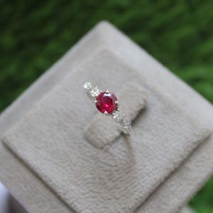 May include: A silver ring featuring a large, round, deep red gemstone as the focal point. Smaller, clear gemstones are set along the band, adding sparkle. The ring is displayed on a light gray surface.