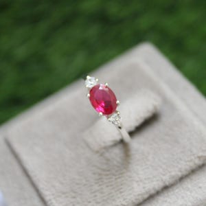 Può includere: Un anello d'argento con una pietra preziosa ovale di rubino rosso e due pietre bianche più piccole su entrambi i lati.