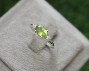 Puede incluir: Un anillo de plata con una piedra preciosa ovalada verde de peridoto y dos piedras redondas transparentes más pequeñas.
