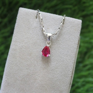 Op de afbeelding: Een zilveren ketting met een druppelvormige, helderrode hanger. De hanger is gezet in een zilveren rand en hangt aan een fijne zilveren ketting. De ketting wordt gepresenteerd op een lichtgrijze ondergrond tegen een vage groene achtergrond.