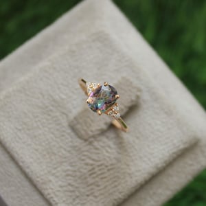 May include: A gold ring featuring an oval-cut, iridescent gemstone as the centerpiece. The ring has small, clear accent stones on either side of the main stone. The ring is set on a neutral-colored surface with a blurred green background.
