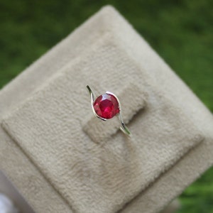 May include: A silver ring with a single, round, red gemstone set in the center. The ring has a curved, twisted band.